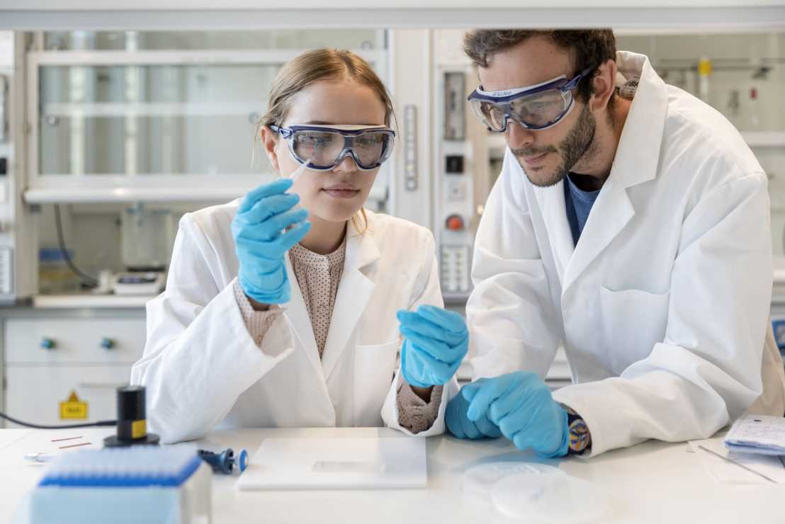 Oksana Dudaryeva and Giovanni Bovone in lab coats and goggles working with a test tube